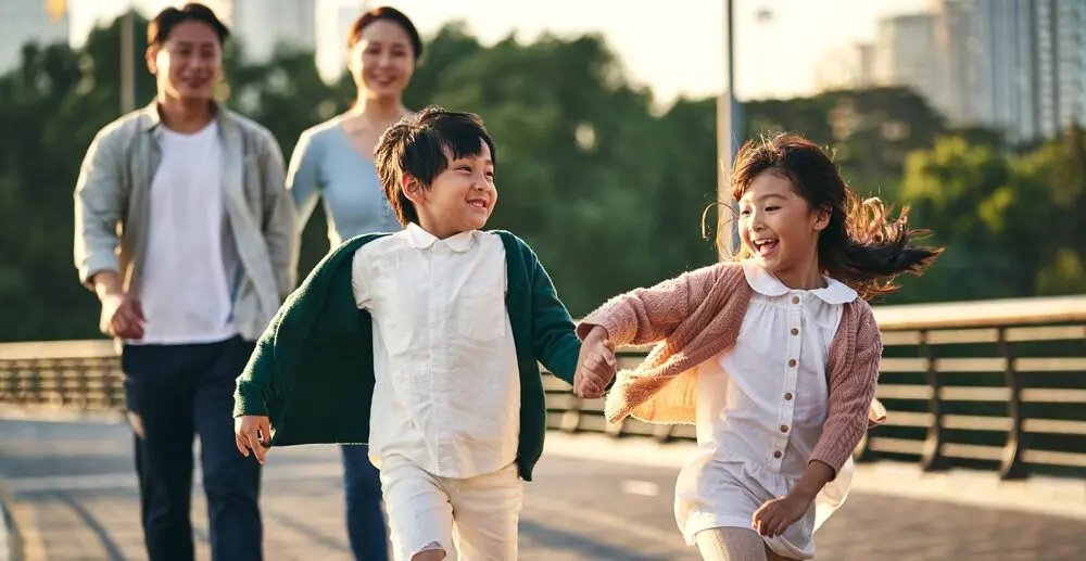 子どもたちが自由にのびのびと育つ未来の象徴 | my stellaマイステラブログ|子どもの好きが育つヒントと親子の時間 笑顔で手をつなぎながら走る男の子と女の子、その後ろを見守る両親の姿