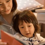 ベッドで母親が子どもに寄り添いながら絵本を読み聞かせている様子