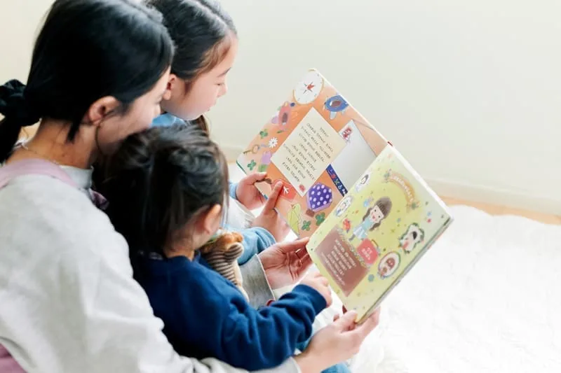 母親と2人の子どもが床に座ってMY STELLAの名前入り絵本を一緒に読んでいる様子。カラフルなイラストが描かれた絵本のページを指差しながら、家族で物語を楽しんでいる読み聞かせタイム。